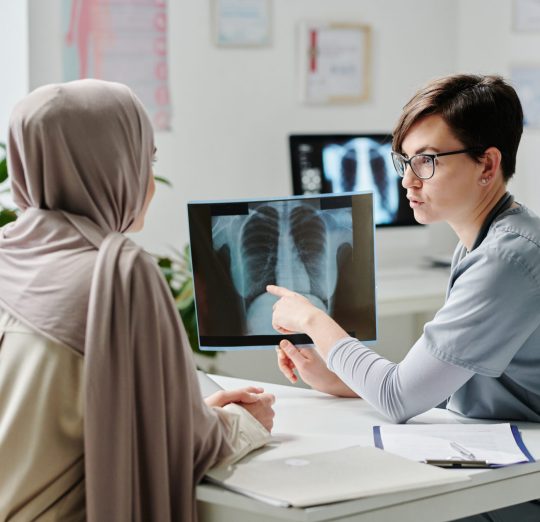 Confident female radiologist showing result of medical examination to patient in hijab while sitting by desk in front of her