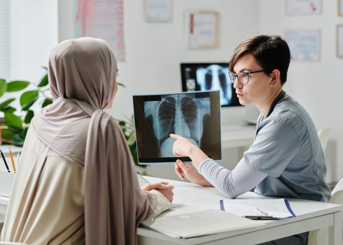 Confident female radiologist showing result of medical examination to patient in hijab while sitting by desk in front of her