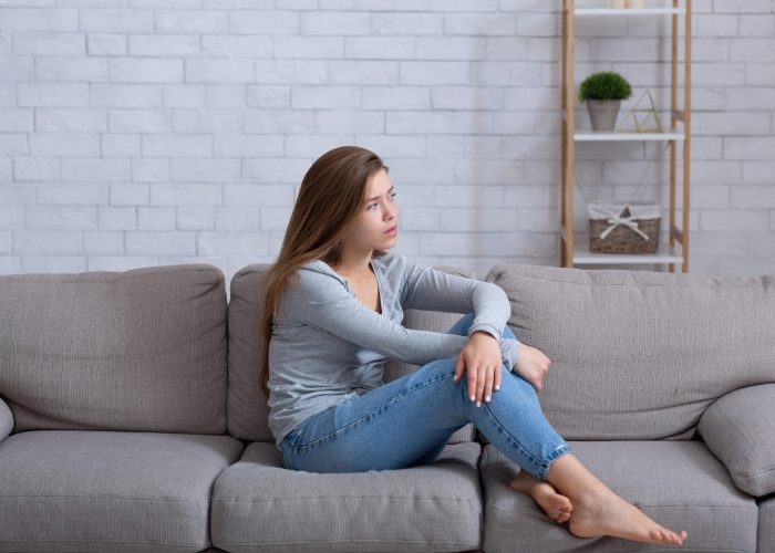 Lonely young lady sitting on sofa at home, feeling depressed and frustrated, suffering from emotional exhaustion. Millennial woman having psychological problem, mental or mood disorder